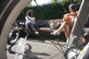 Looking through the spokes of a bicycle wheel we see two women chatting on a bench